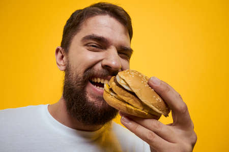 Man with hamburger in the hands of fast food diet food white t-shirt close-up yellow background