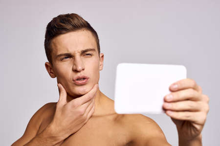 A half-naked man with a mirror in his hand examines a cropped view with a face on a light background