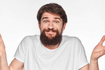 cheerful bearded man in a white t-shirt emotions gestures with his hands light background studio