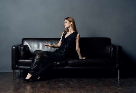 Woman in black dress sits on leather sofa indoors evening makeup naked shoulders model