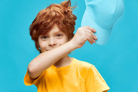 Red-haired boy with freckles holds a cap in his hands in a yellow t-shirt blue isolated fundの写真素材
