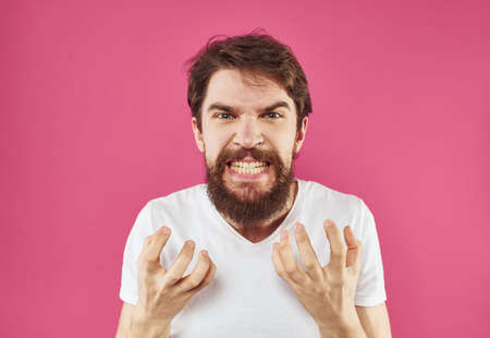 A man with a beard gestures with his hands near his face on a pink background