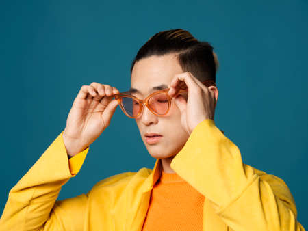 Asian man straightens glasses on his face and yellow jacket blue backgroundの写真素材