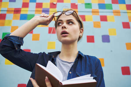 A student in a shirt and glasses holds a book in her hands on the street near the graffiti on the...