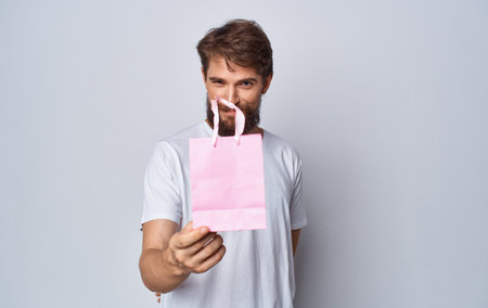 bearded man in white t-shirt holding pink gift bag in hands isolated backgroundの写真素材