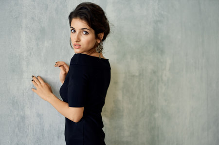 romantic brunette in an evening dress leaning against a gray wall indoors earrings makeup modelの写真素材