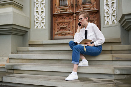 female student sits on the steps near the building with a book in her hands and a mobile phone