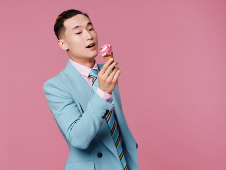 A man of Asian appearance eats ice cream in a blue suit pink backgroundの写真素材