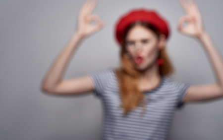 Emotional woman in glasses and in a striped T-shirt gestures with her hands red hat gray background