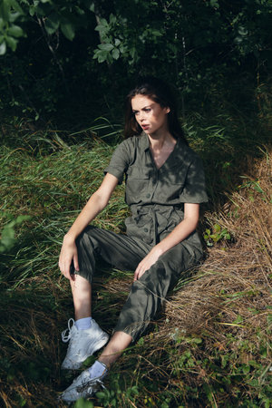 A woman in sneakers and overalls sits on dry grass in the forestの写真素材