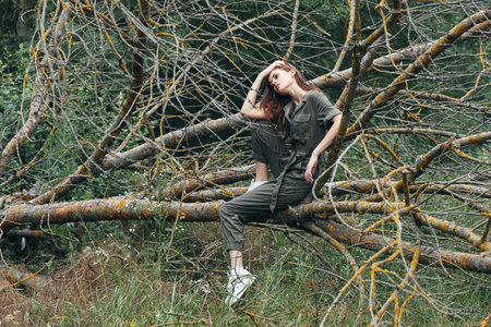 A woman in a green jumpsuit touches her head and sits on a broken tree