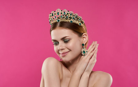 A romantic princess with a diadem on her head touches her face with her hands on a pink backgroundの写真素材