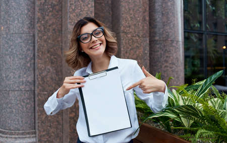 Happy business woman with documents near the building on the street in glasses and in a white shirt