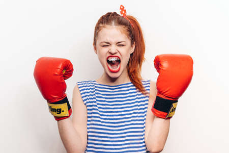 red-haired woman in boxing gloves and a striped T-shirt on a light background with an open mouthの写真素材