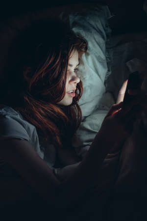emotional woman with a phone in her hands lies in bed at night addictionの写真素材