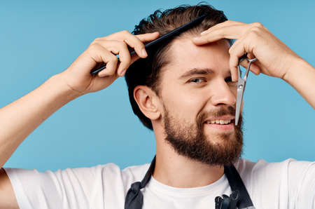 a man hairdresser in a gray apron does his hair on a blue background scissors comb