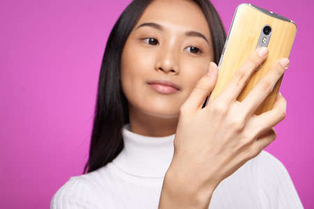 pretty asian woman with a phone in her hands looking at the screen close-up pink background
