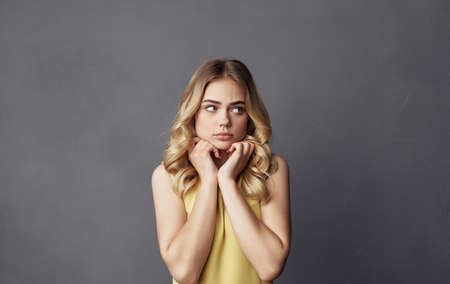 cheerful pretty blonde yellow dress emotions hands near face cropped view gray background