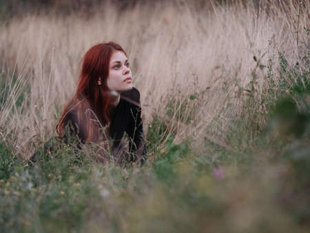 red-haired woman in a black dress lies in a field on dry grass in nature