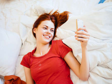 Happy woman lies in bed and looks at the screen of a mobile phone