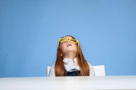 emotional schoolgirl sitting at the table yellow glasses in hands blue backgroundの写真素材