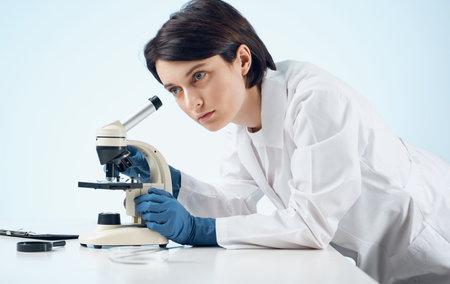 woman doctor in a medical gown and gloves looking through a microscope on a table indoors