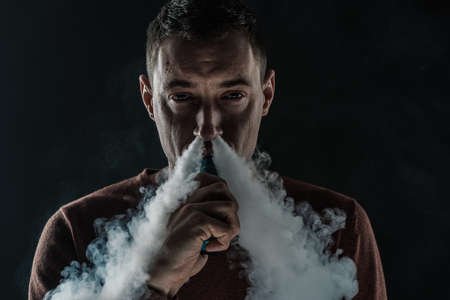a man smokes a vape on a black background white smoke steam portrait close-upの写真素材