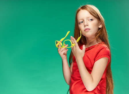 girl with red hair holding yellow glasses in her hands green background studio red t-shirt