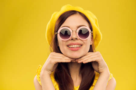 Happy woman in a yellow hat and fashionable glasses holds her hands near her face on a yellow background cropped view of emotions funの写真素材