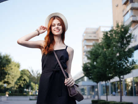 Pretty red-haired woman in a black dress outdoors on the street near the buildingの写真素材