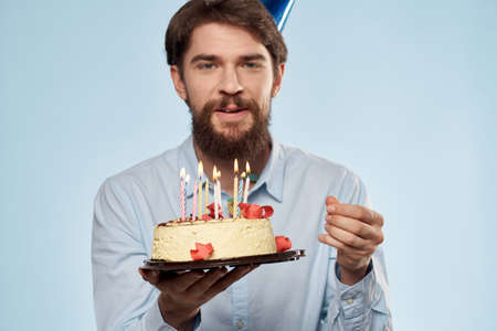 Bearded man with a plate of cake on a blue background and a birthday party hat on his head