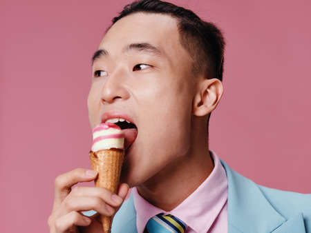 Happy Asian man eating ice cream on pink background in blue classic suit cropped view