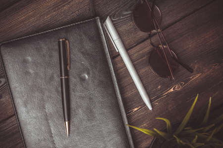 book with black cover and lies on a wooden table and a pen pencil jobs glasses flower potted coffee cupの写真素材