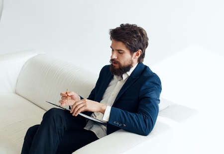 A business man in a suit sits on a white sofa In a bright room, side view