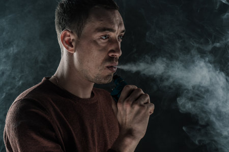 a man smokes a vape on a black background white smoke steam portrait close-upの写真素材