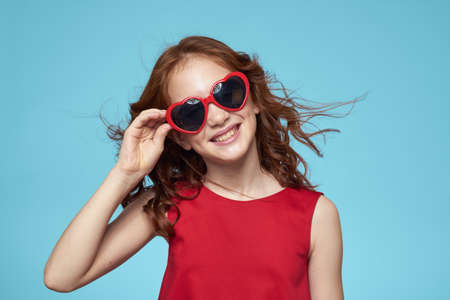 Cheerful little girl with sunglasses in the form of a heart red dress blue backgroundの写真素材