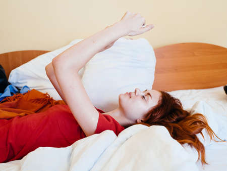 Happy woman with mobile phone lies in bed and looks at the screenの写真素材