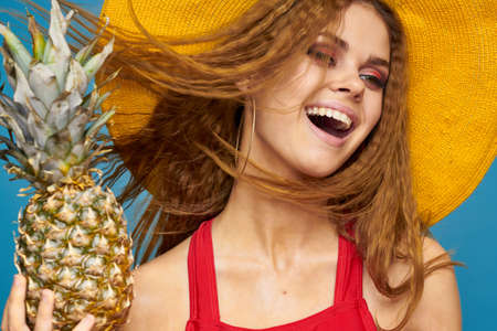 Woman in yellow hat with pineapple hands curly hair exotic fruits blue background attractive lookの写真素材