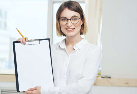 A woman is holding a folder with a white sheet of mockup paper and a window in the backgroundの写真素材