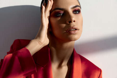 beautiful woman with evening make-up and in a red jacket looks to the side and holds her hand near her faceの写真素材