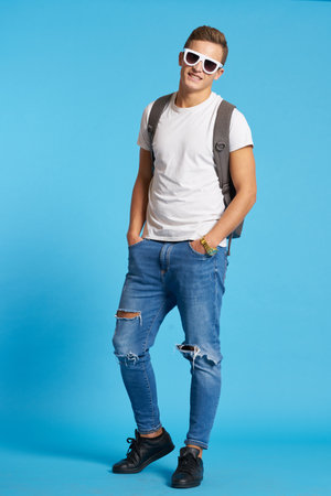 A man with a backpack in glasses and in jeans sneakers T-shirt blue background in full growthの写真素材