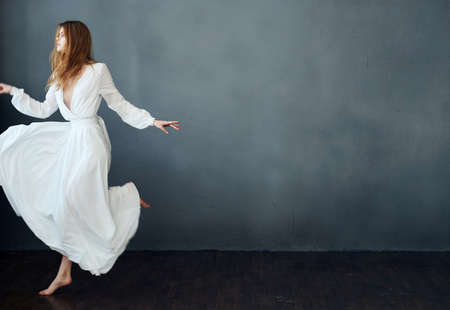 A woman in a white dress is dancing on a gray background in full growth red hair modelの写真素材