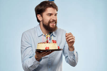 Bearded man with a plate of cake on a blue background and a birthday party hat on his headの写真素材