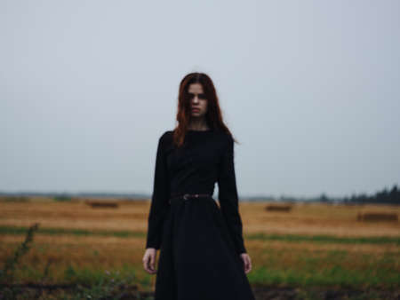 Beautiful woman with red hair in a black dress outdoors in the fieldの写真素材