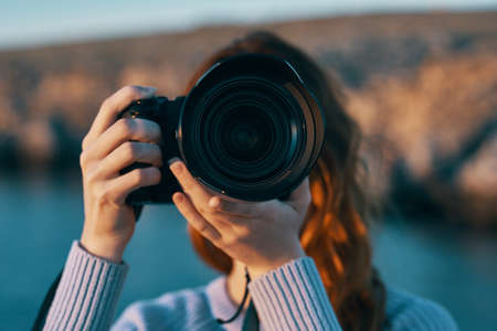 woman holding a camera with a lens in nature in the mountains and the sea in the backgroundの写真素材