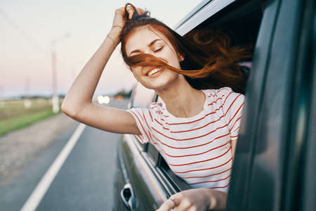 energetic red-haired t-shirt woman peeking out of the open car window on the road track journeyの写真素材