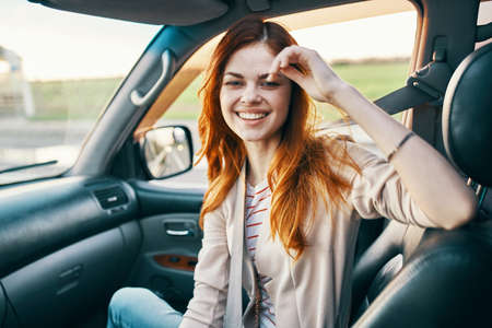 happy red-haired woman in a beige coat in the front seat of a car smile model gesturing with her...