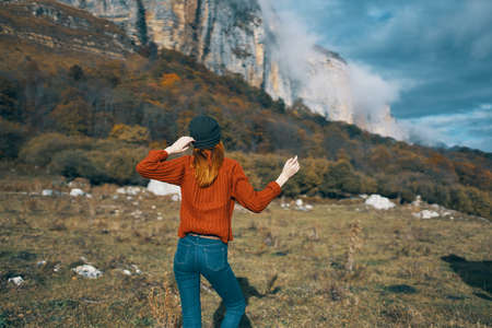 a woman in a sweater holds her hands behind her head and looks at the high mountains landscape blue...
