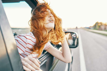 happy red-haired woman looks out of the car window and drives on the road, track summer vacationの写真素材