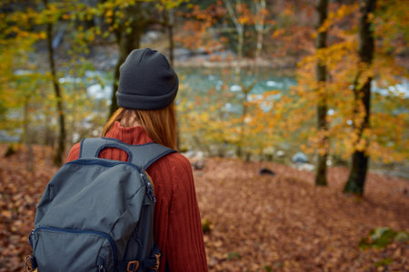 woman in a sweater with a backpack on her back near the river in the mountains and park trees autumn...
